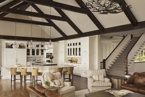 great room kitchen, white kitchen, beaned ceiling, wood floor, island stools, two islands, pendant lights, glass front cabinets, armchairs, stairway, ship lap, chandelier, counter stools, arm chairs, pillows, medium wood floor, beamed ceiling, glass-fronted cabinets, two islands