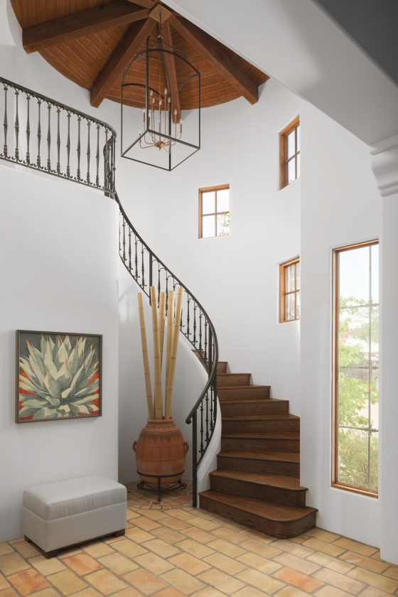 curving stairway, terra cotta tile floor, bench, wall art, clay pot, bamboo branches, large ceiling fixture, wood ceiling, mullioned windows, white walls, wrought iron staircase railing, wood stairs