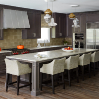kitchen, medium brown cabinets, medium brown wood floor, medium brown island with light countertop, white counter stools with stained wood legs, subway tile backsplash, pendant lights