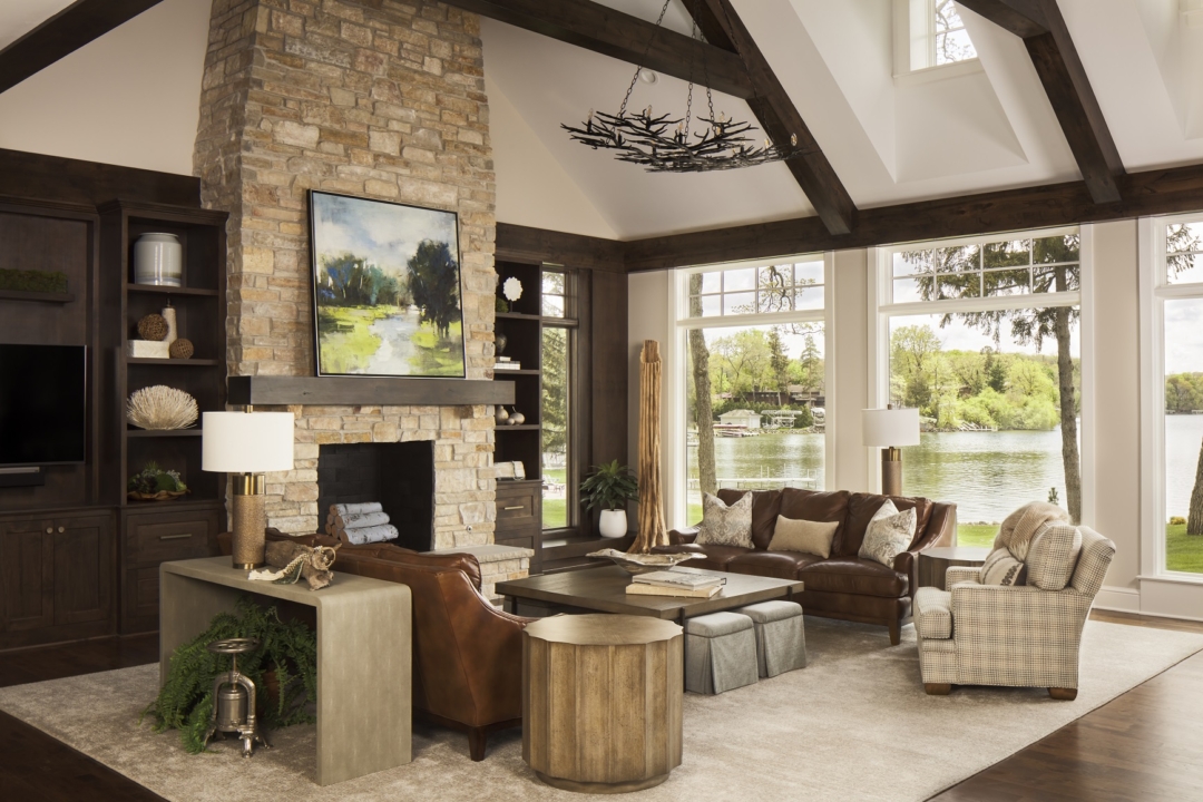 black metal branch chandelier, lakeside great room, stone fireplace, brown sofas, grey ottomans, soft white rug, grey and cream arm chair, light wood console, table lamp, round gear-like side table, wall art, windows with lake view